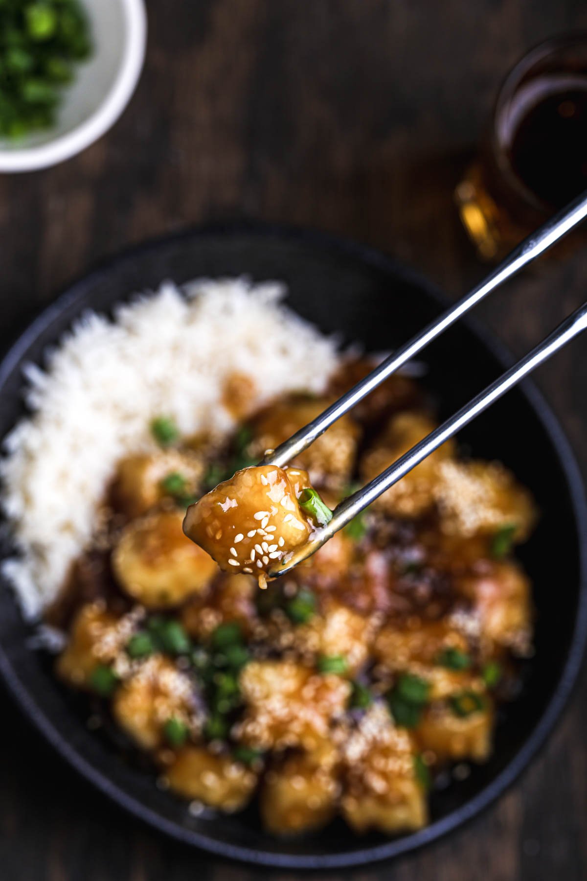 Chopsticks lifting a piece of General Tso’s tofu coated in sticky sauce with sesame seeds.