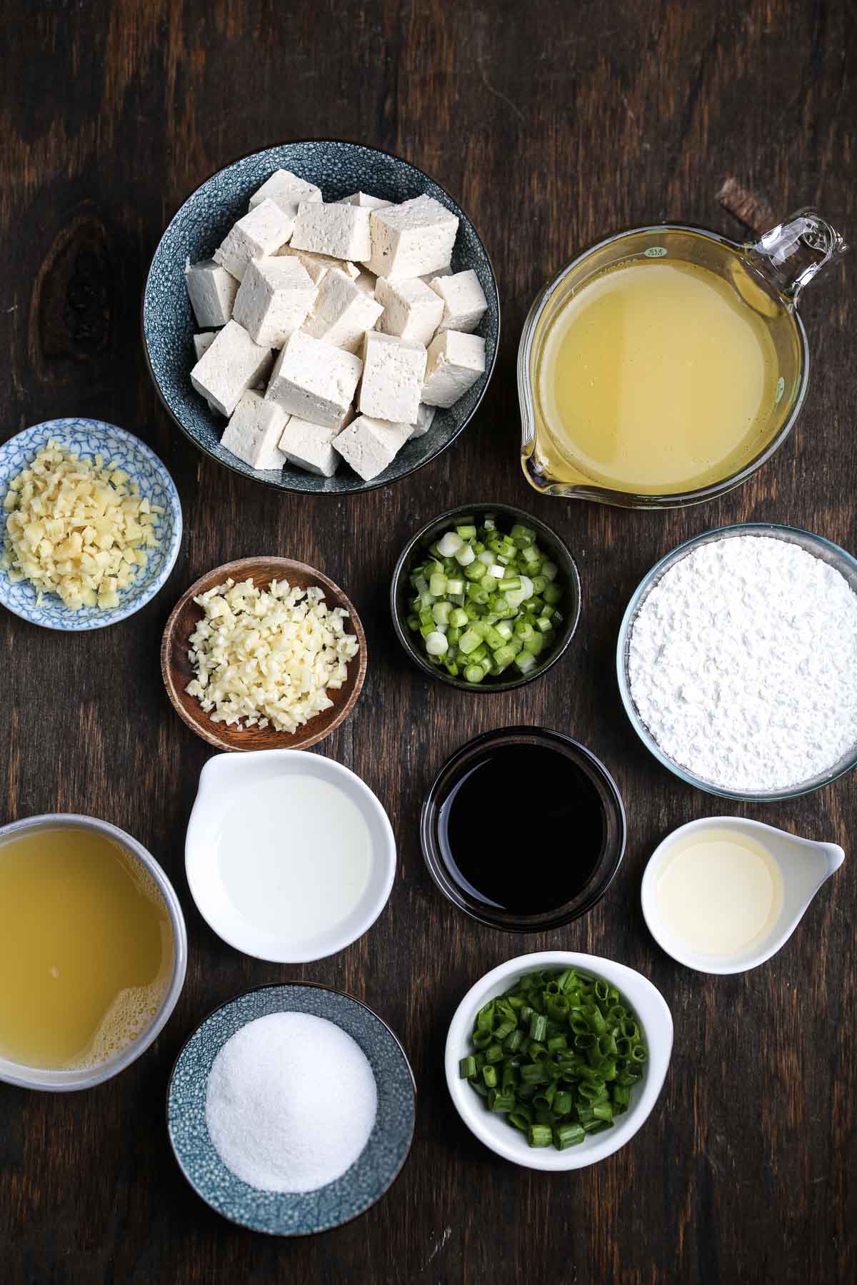 Ingredients for General Tso’s tofu laid out on a wooden table, including tofu, scallions, garlic, soy sauce, and cornstarch.