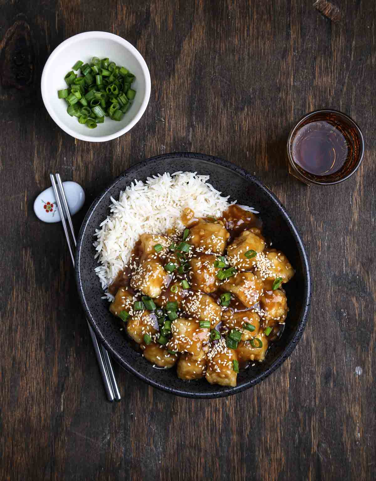 General Tso’s tofu served with steamed rice, scallions, and sesame in a black bowl.