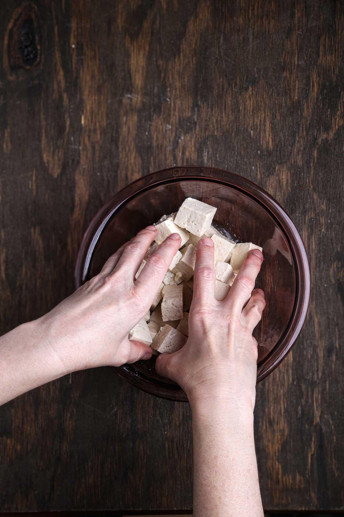 Hands tossing cubed tofu in a glass bowl before dredging.