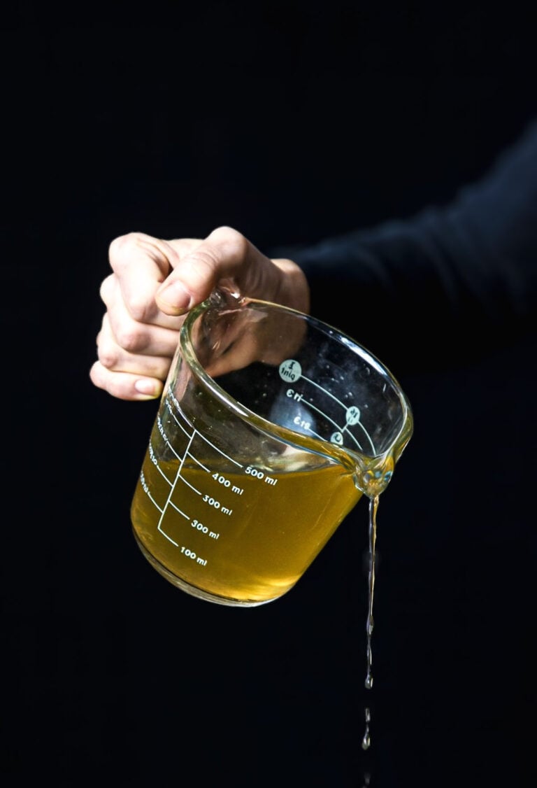 Hand pouring golden vegan stock from a glass measuring cup against a black background.