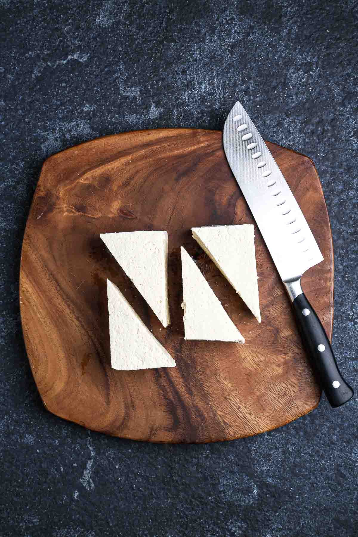 Firm tofu cut into four triangles on a wooden cutting board with a chef’s knife ready for frying.
