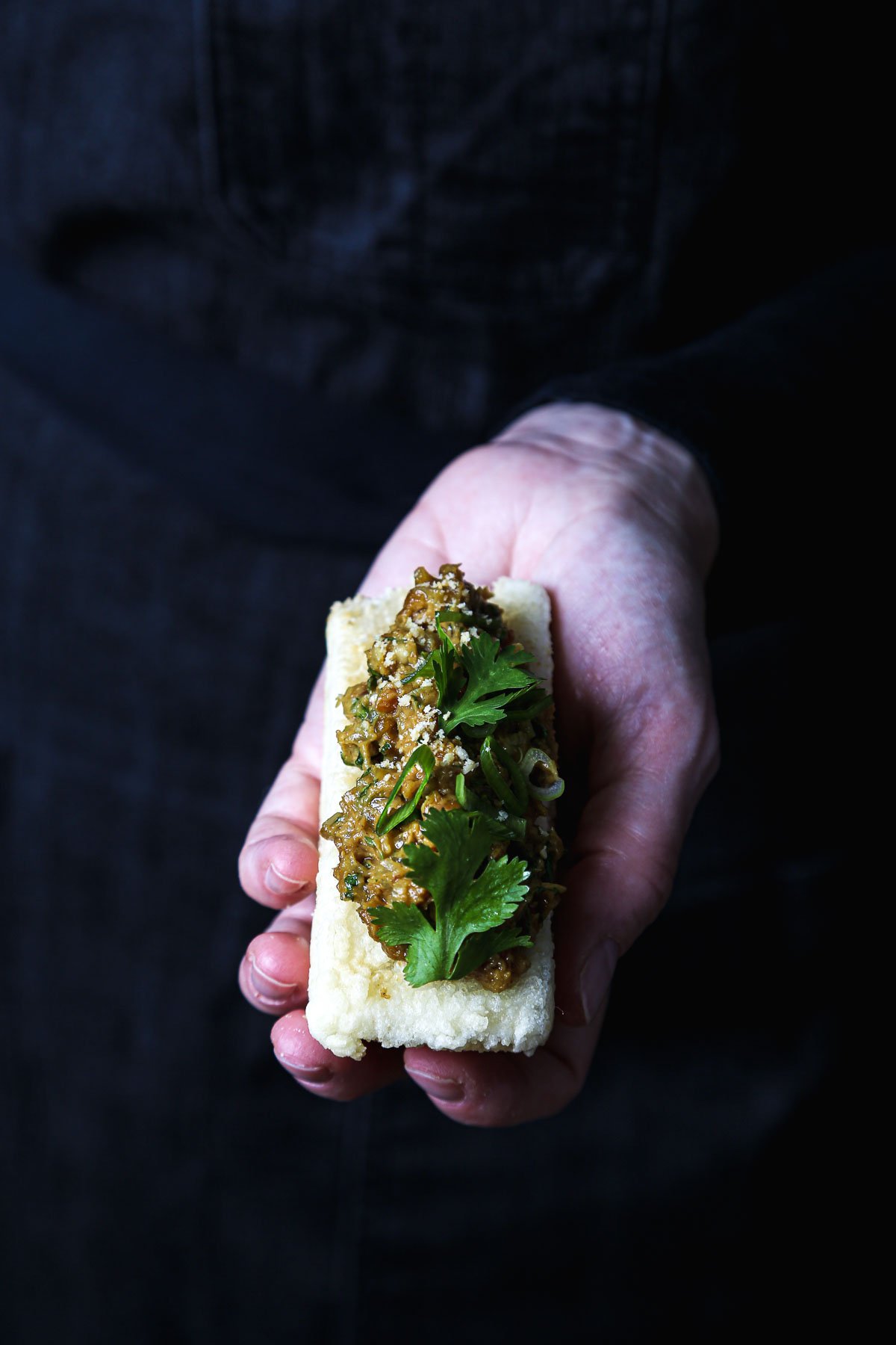 Hand holding a piece of crispy fried tofu filled with lemongrass–peanut sauce, topped with cilantro and scallions against a dark background.