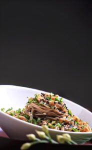 Bowl of soba noodles with sesame-ginger sauce, black sesame seeds, and fresh herbs, set against a dark background.