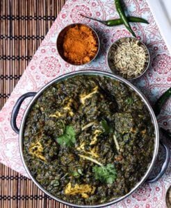 Overhead view of vegan palak paneer served in a metal bowl, surrounded by fennel seeds, curry powder, and green chiles.