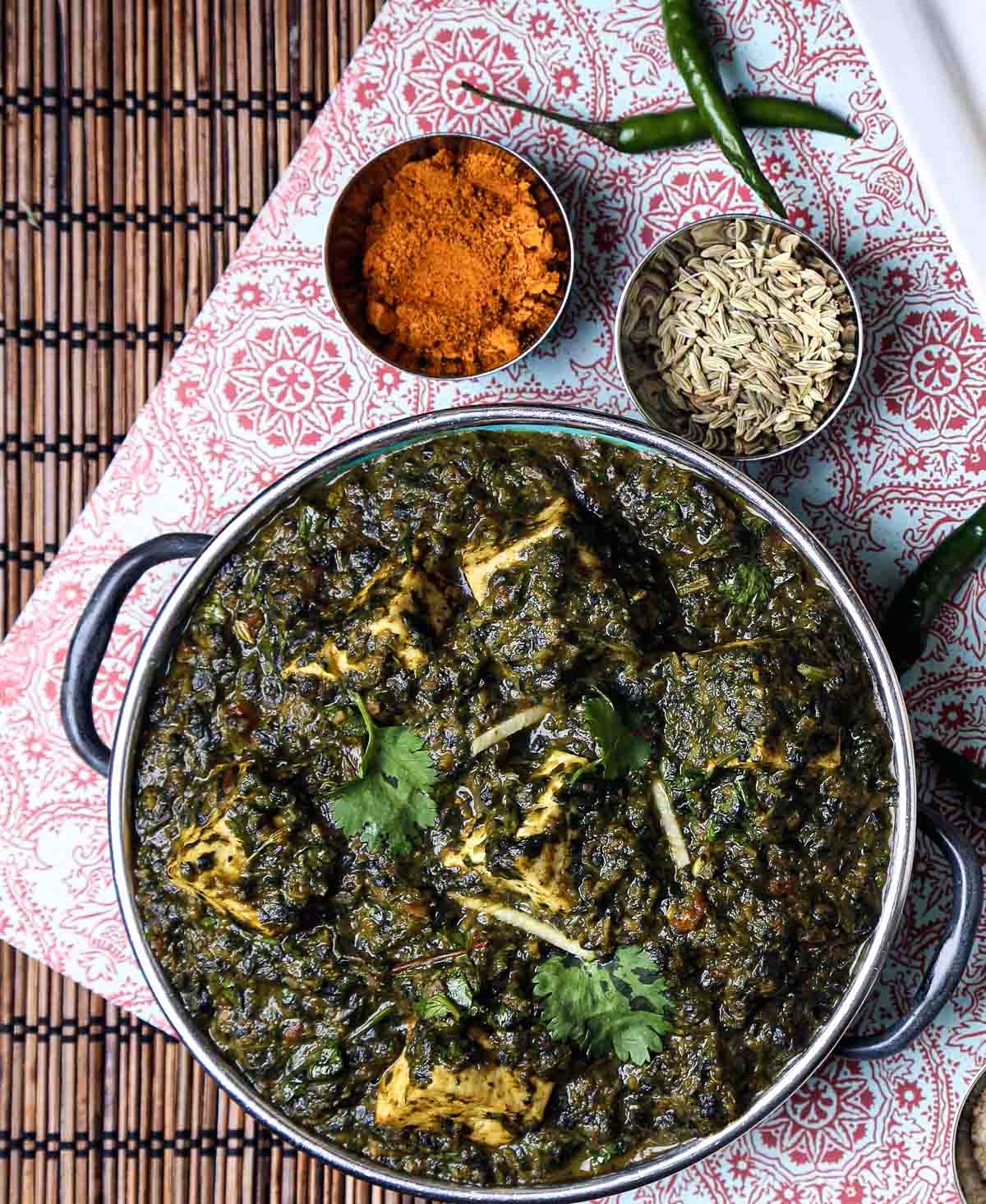 Overhead view of vegan palak paneer served in a metal bowl, surrounded by fennel seeds, curry powder, and green chiles.