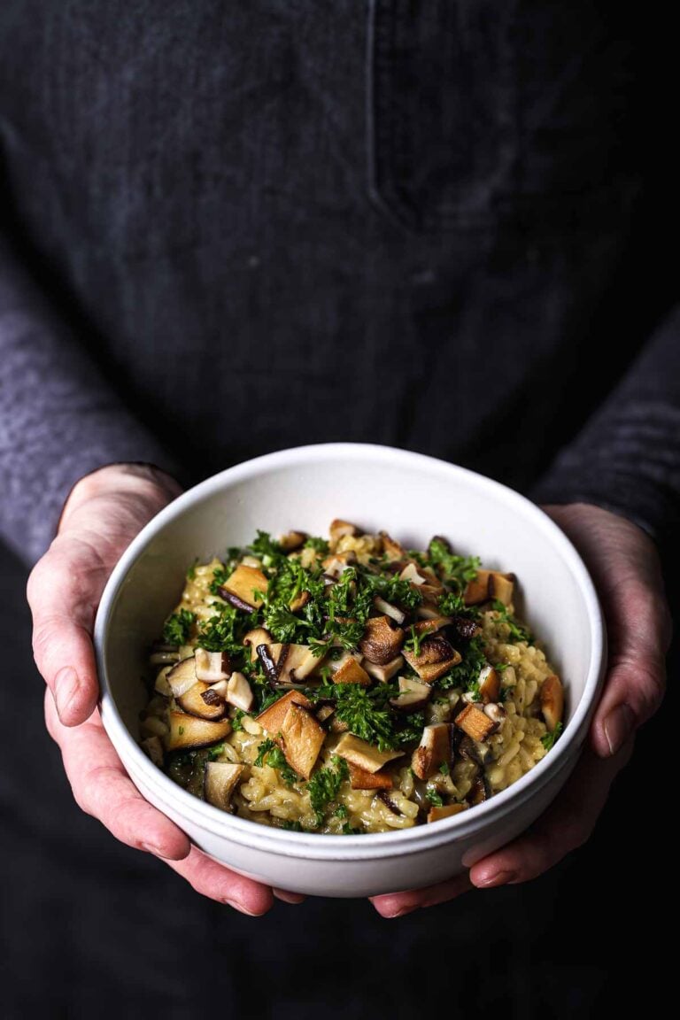 Vegan mushroom risotto topped with seared shiitakes and fresh herbs, served in a white bowl.