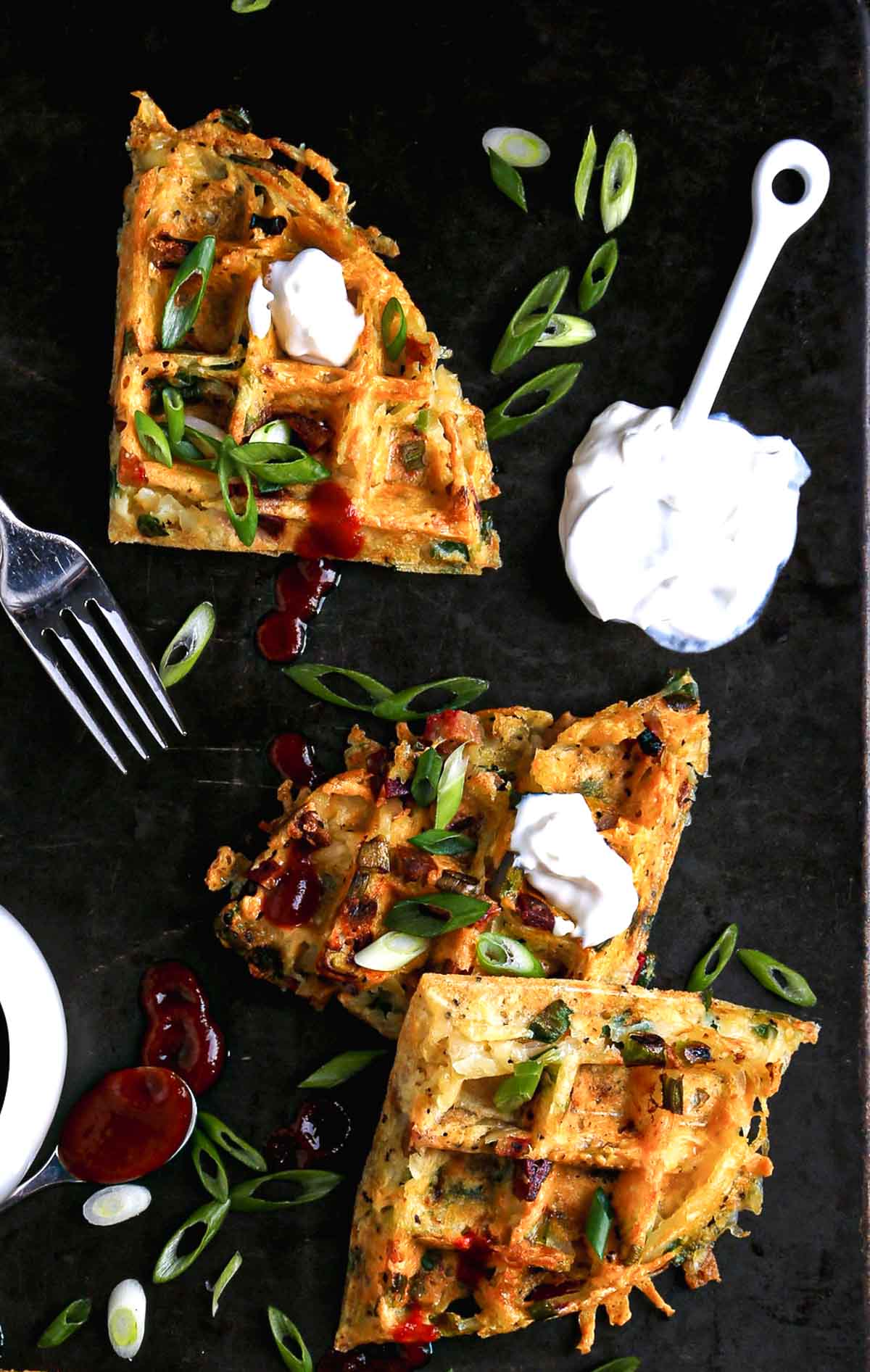 Golden, crispy hash brown waffles topped with vegan sour cream, scallions, and sriracha on a dark plate.
