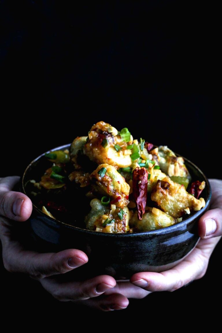 Bowl of crispy kung pao Brussels sprouts held in both hands, coated in a sticky, spicy-sweet glaze with peanuts and scallions.