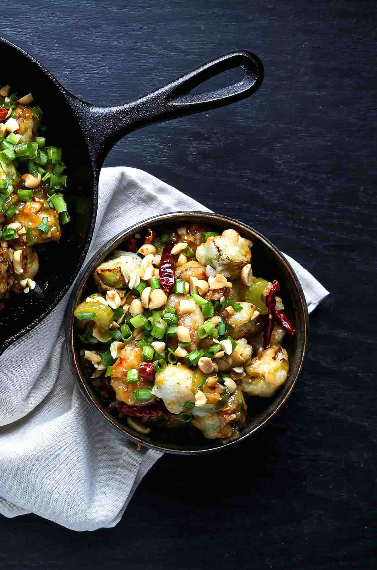 A small bowl of crispy kung pao Brussels sprouts with scallions and peanuts, styled next to a cast-iron skillet.