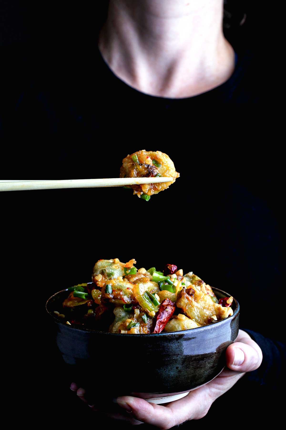 Close-up of a crispy Brussels sprout coated in kung pao sauce lifted with chopsticks.