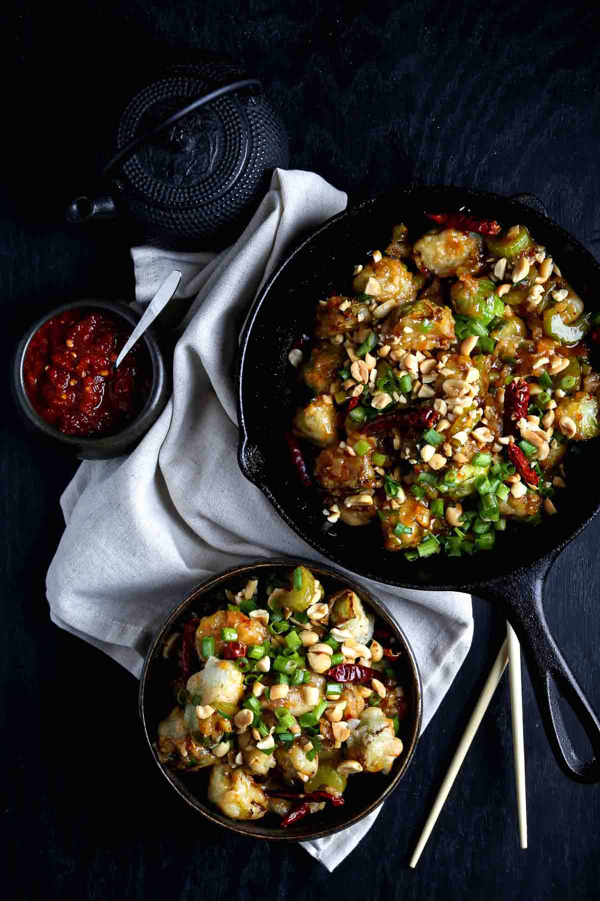 Cast-iron skillet filled with kung pao Brussels sprouts topped with peanuts and scallions, served alongside a small bowl of sambal.