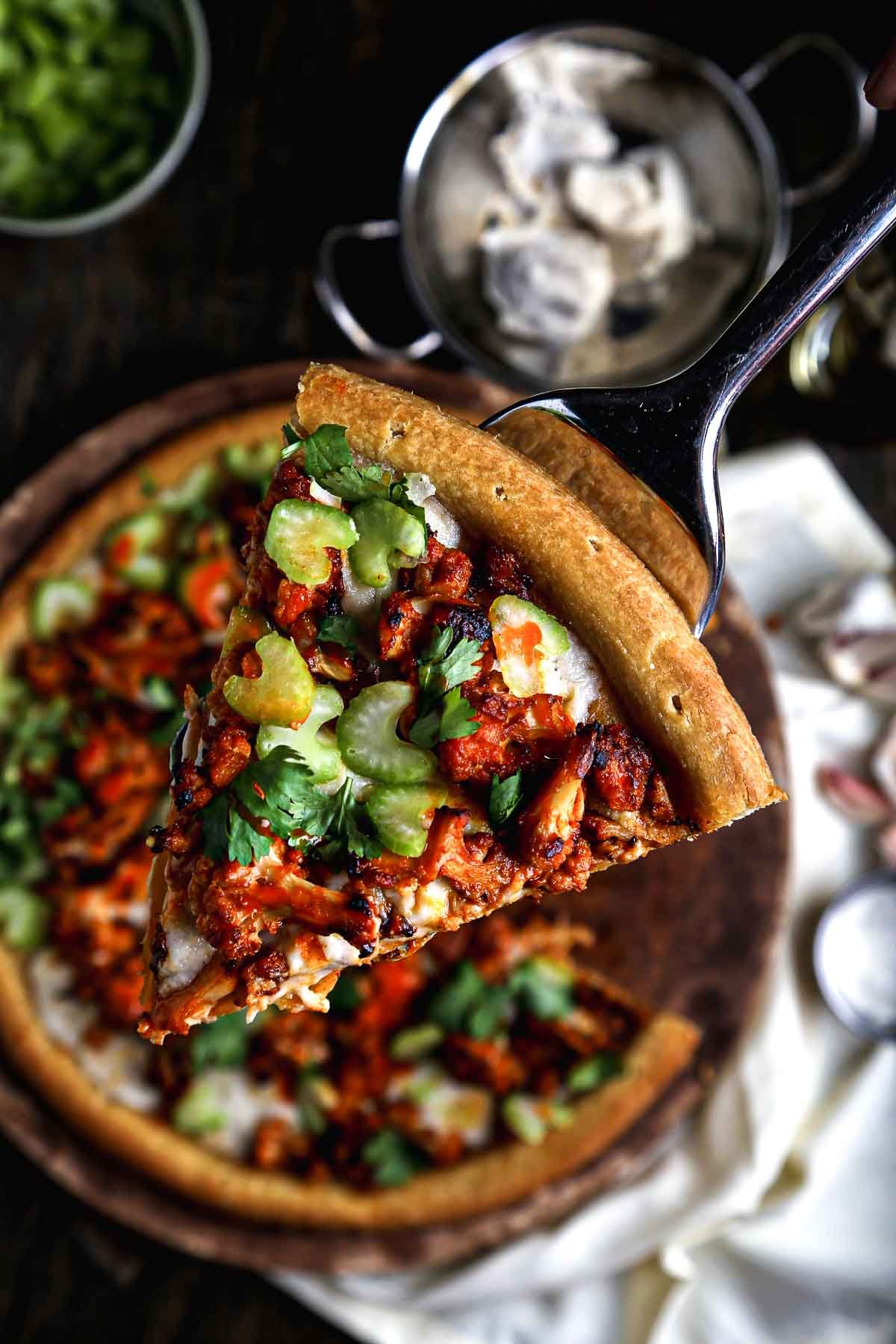 Close-up of a buffalo cauliflower pizza slice with golden crust, vegan cheese, spicy cauliflower, and chopped celery.