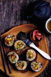 Sweet potato sushi roll with avocado and teriyaki glaze on wooden platter, served with chopsticks, pickled ginger, and tea.