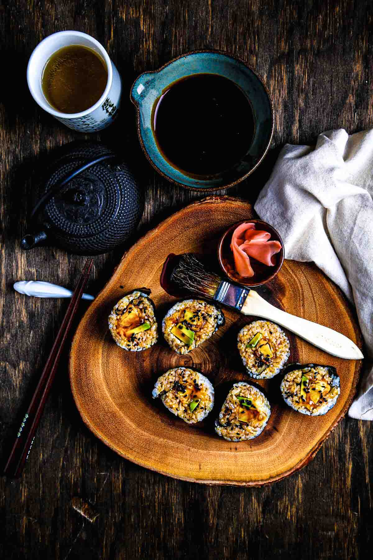 Overhead shot of sushi platter on rustic wood board with dipping sauce, pink pickled ginger, and a cup of green tea.