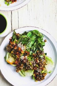 Grilled bok choy topped with oyster mushrooms, chili crisp, and sesame seeds on a white plate.