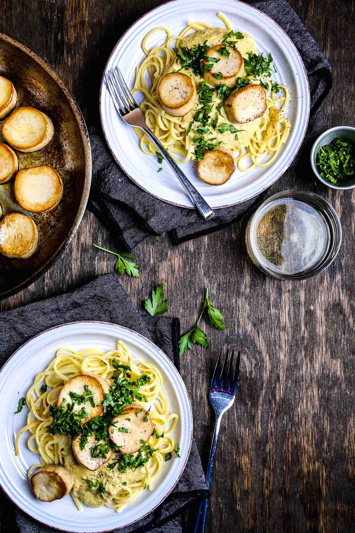 Two plates of creamy vegan mushroom pasta with seared king oyster scallops, parsley, and white wine on a rustic wood table.