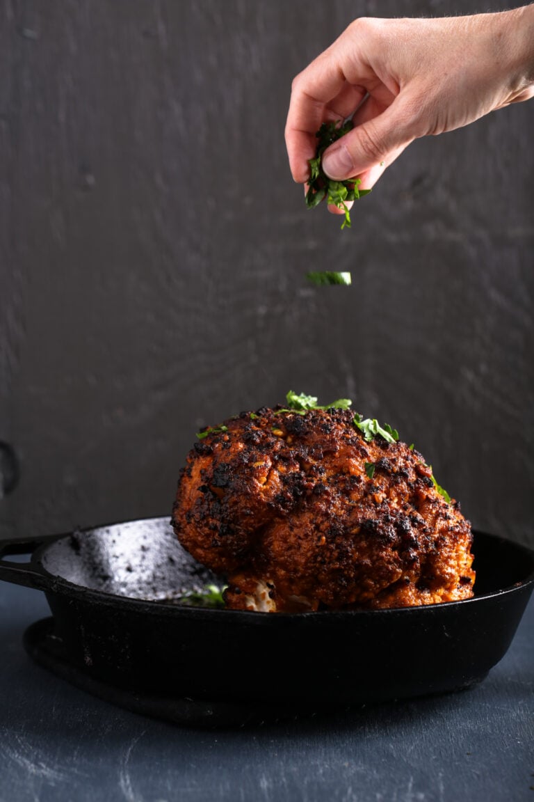 A hand sprinkling parsley over a head of Firecracker Cauliflower in a cast iron pan.