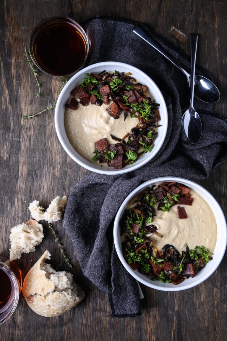 Two bowls of creamy roasted parsnip soup on a wooden board with spoons and bread.