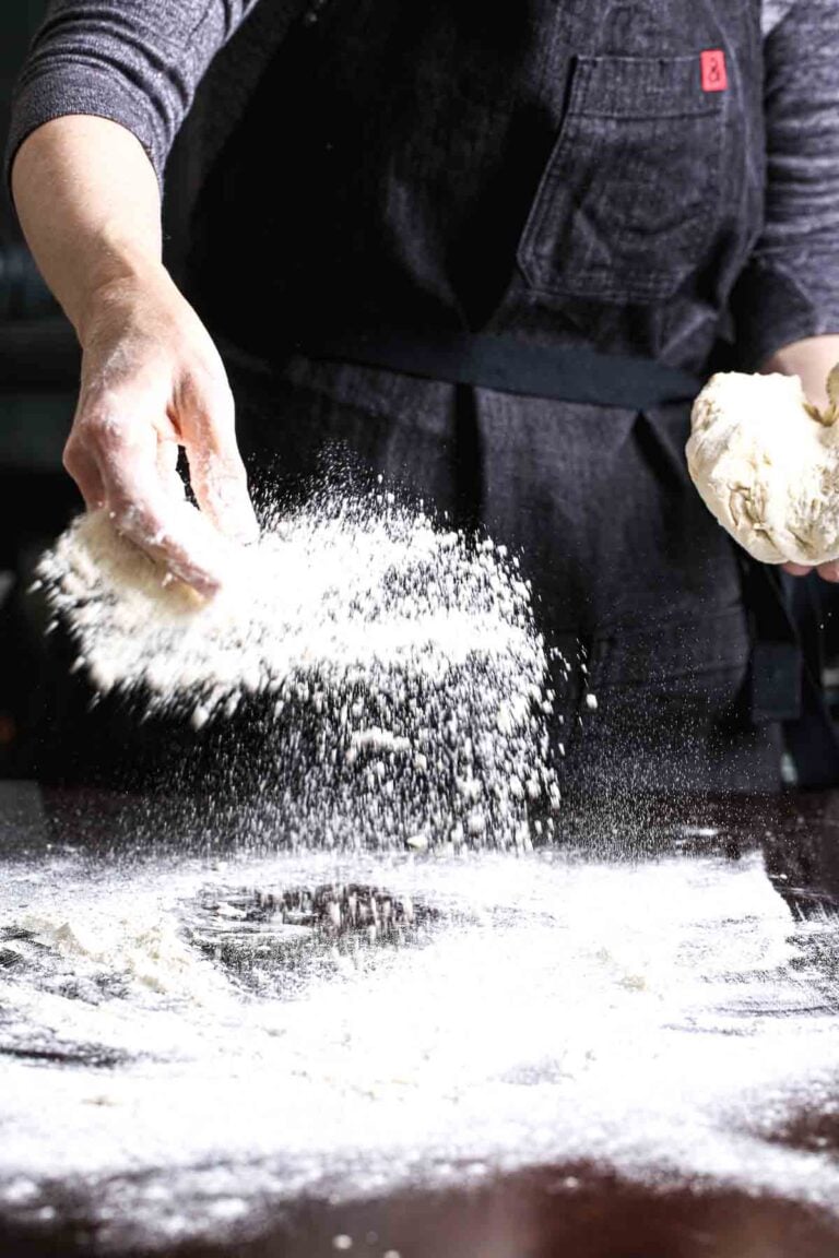 Sprinkling flour over a tabletop to knead vegan pizza dough.