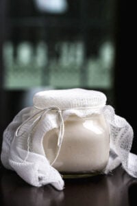 A jar of homemade vegan yogurt covered with cheesecloth, sitting on a dark surface.