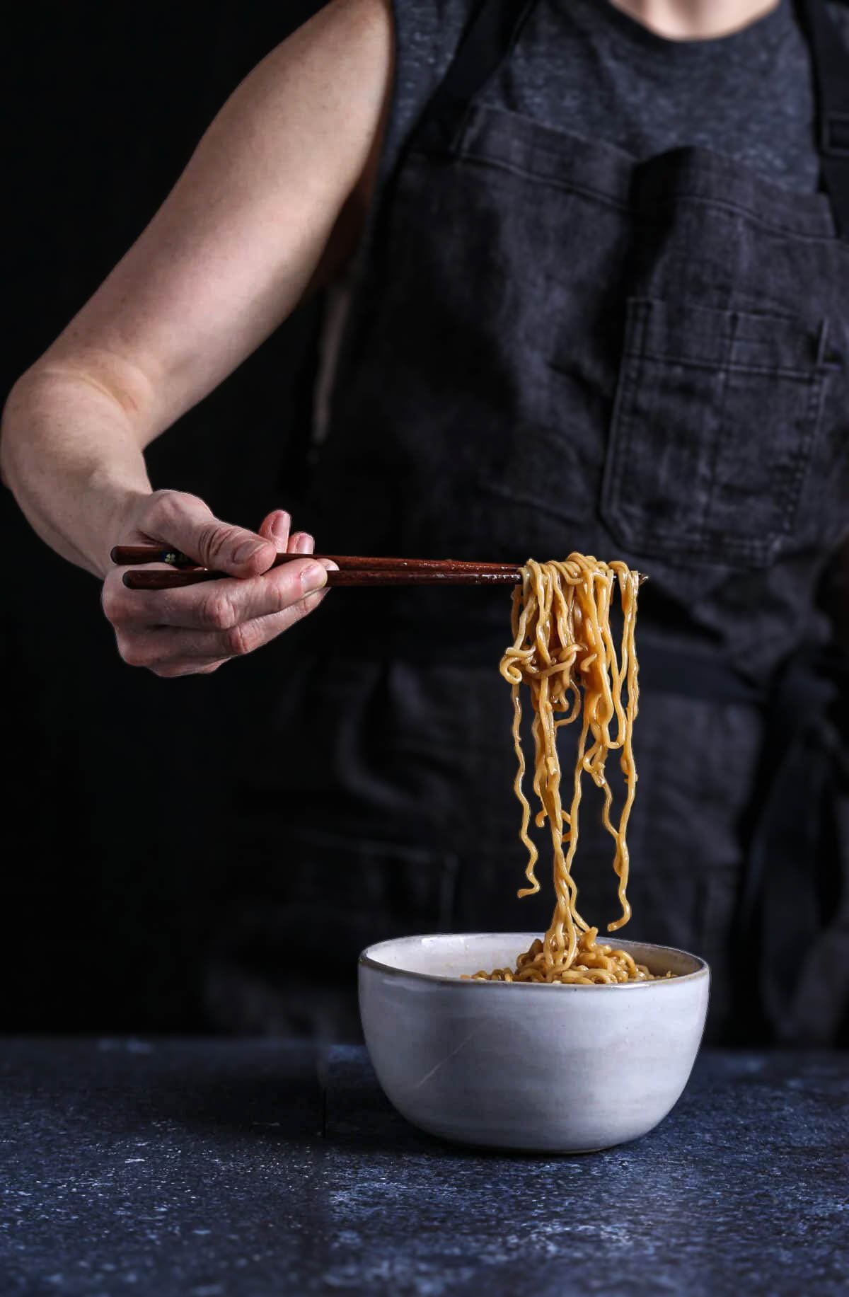 Person lifting ramen noodles with chopsticks over a bowl of umami-rich broth.