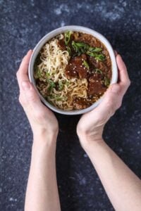 Two hands holding a bowl of vegan French onion ramen with noodles, seared seitan, and scallions.