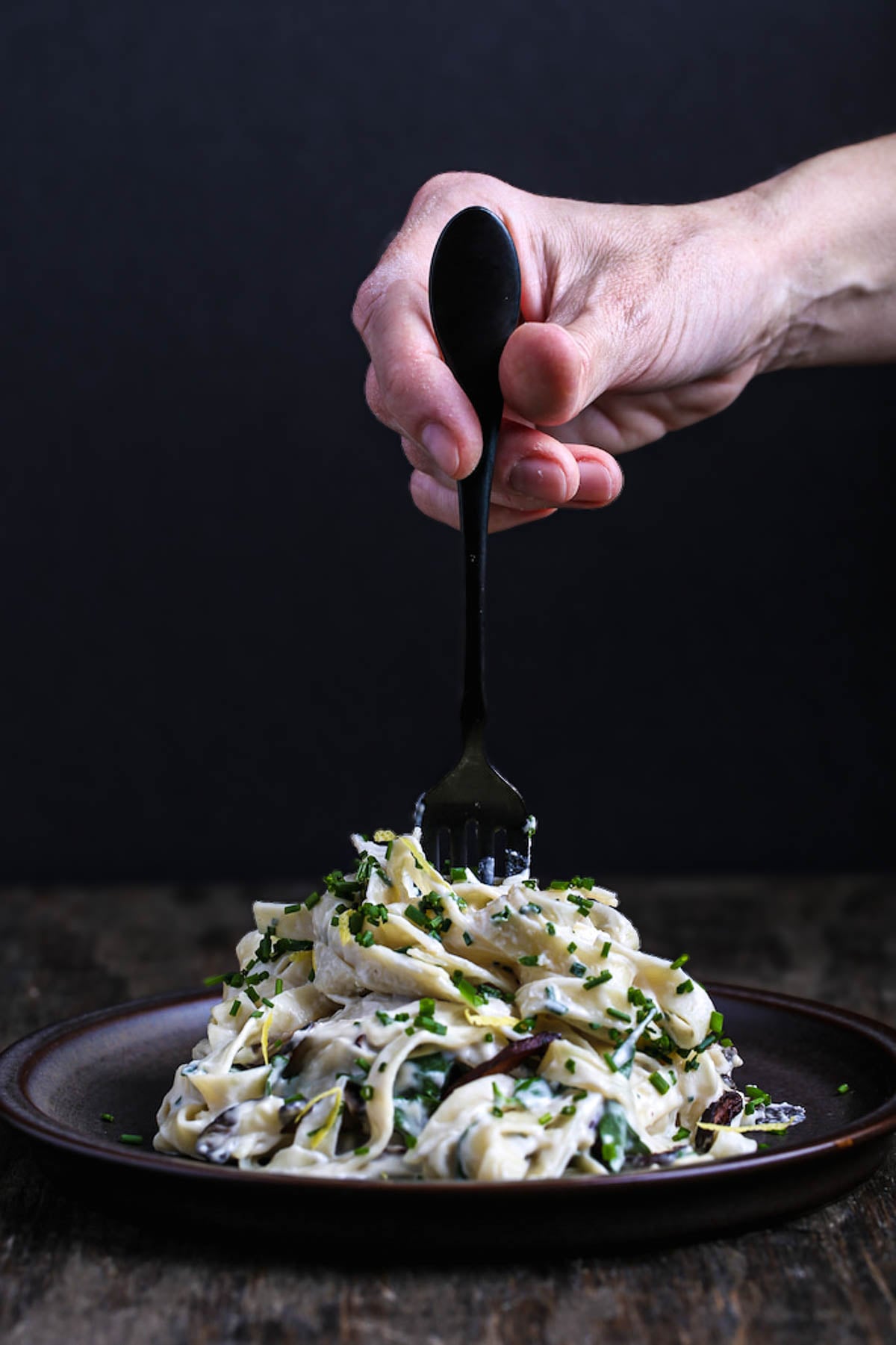 A hand holding a black fork, twirling fettuccine covered in rich vegan alfredo sauce, with chives and shiitake bacon adding the perfect finishing touch.