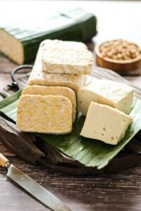 Sliced tempeh and firm tofu blocks arranged on a banana leaf on a wooden board, showing the texture and appearance of two of the best vegan protein sources.