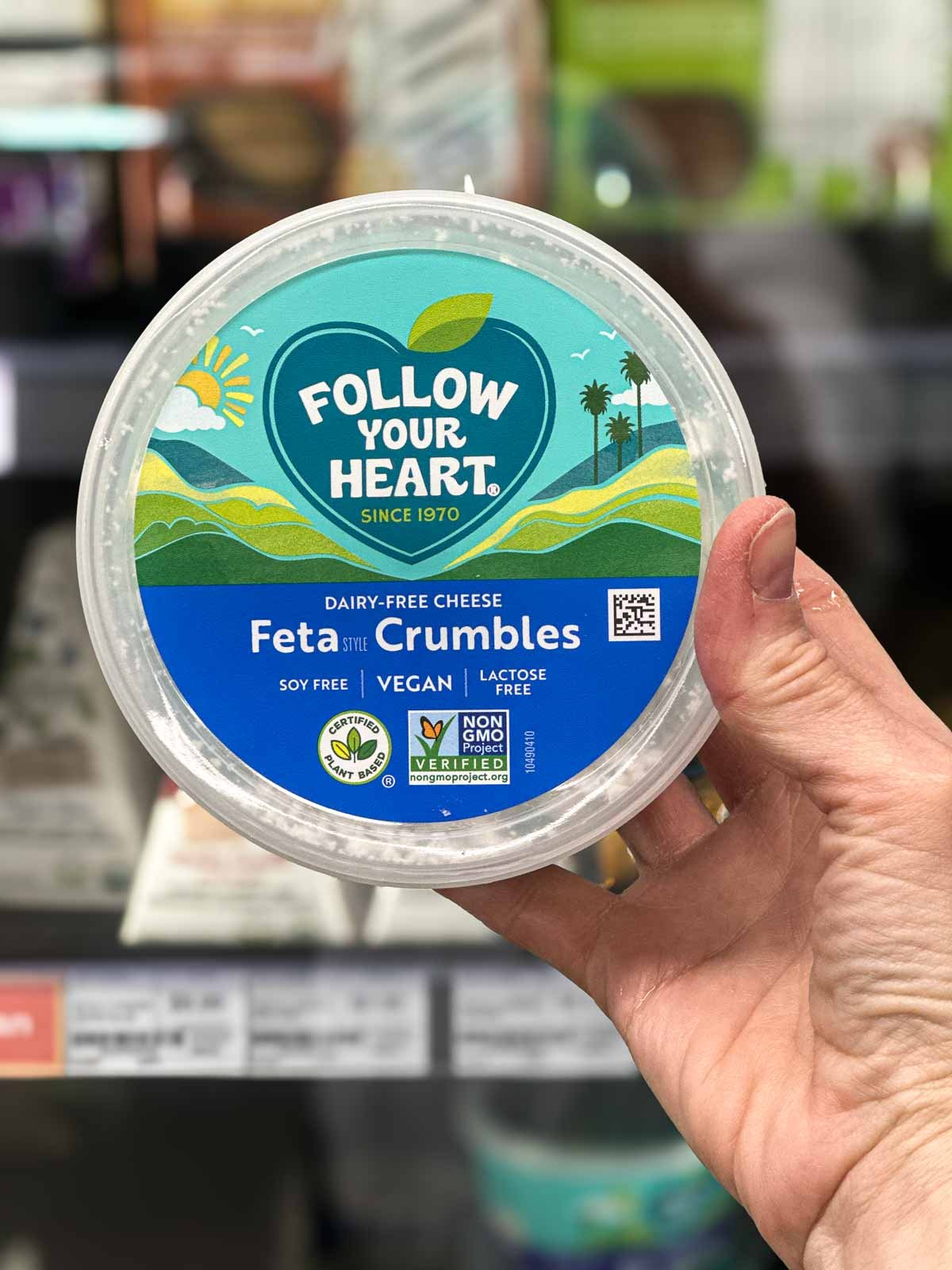 Hand holding a tub of Follow Your Heart plant-based feta cheese crumbles in front of a grocery store dairy case.