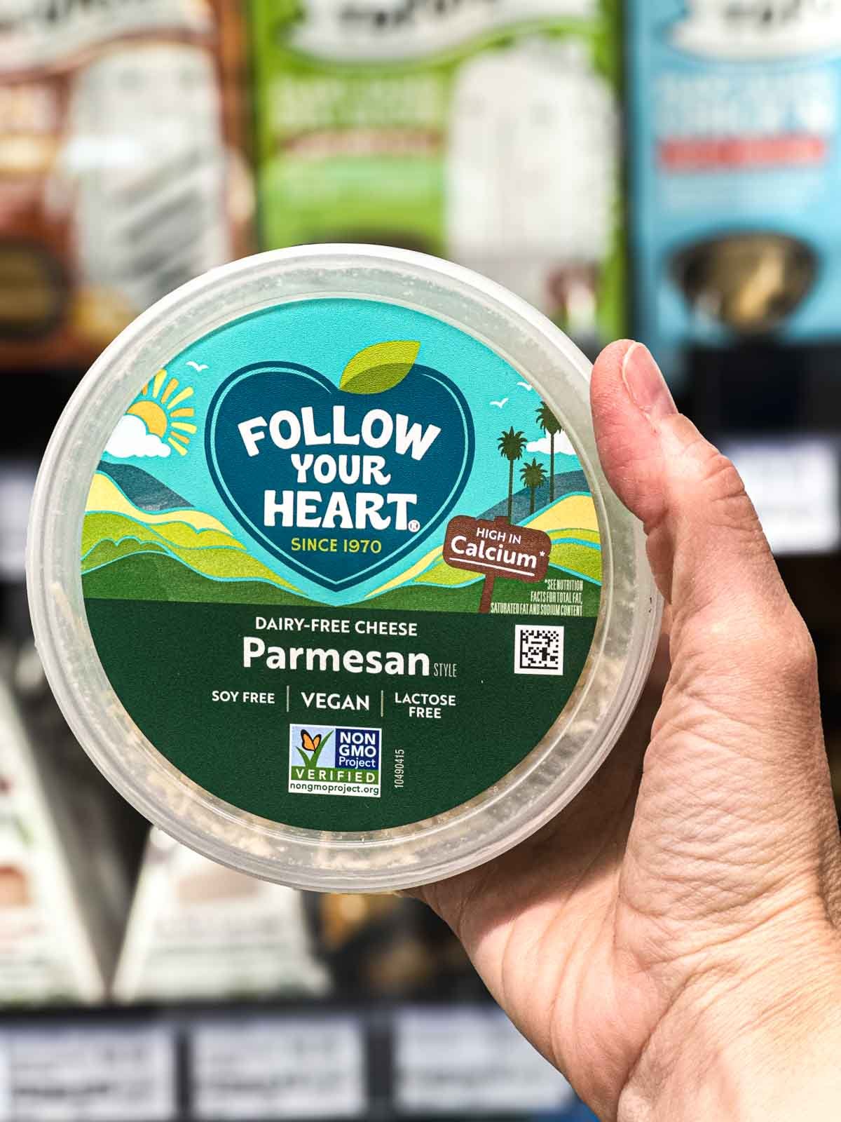 Follow Your Heart dairy-free parmesan cheese in a green-labeled tub, held up in the vegan section of the fridge aisle.