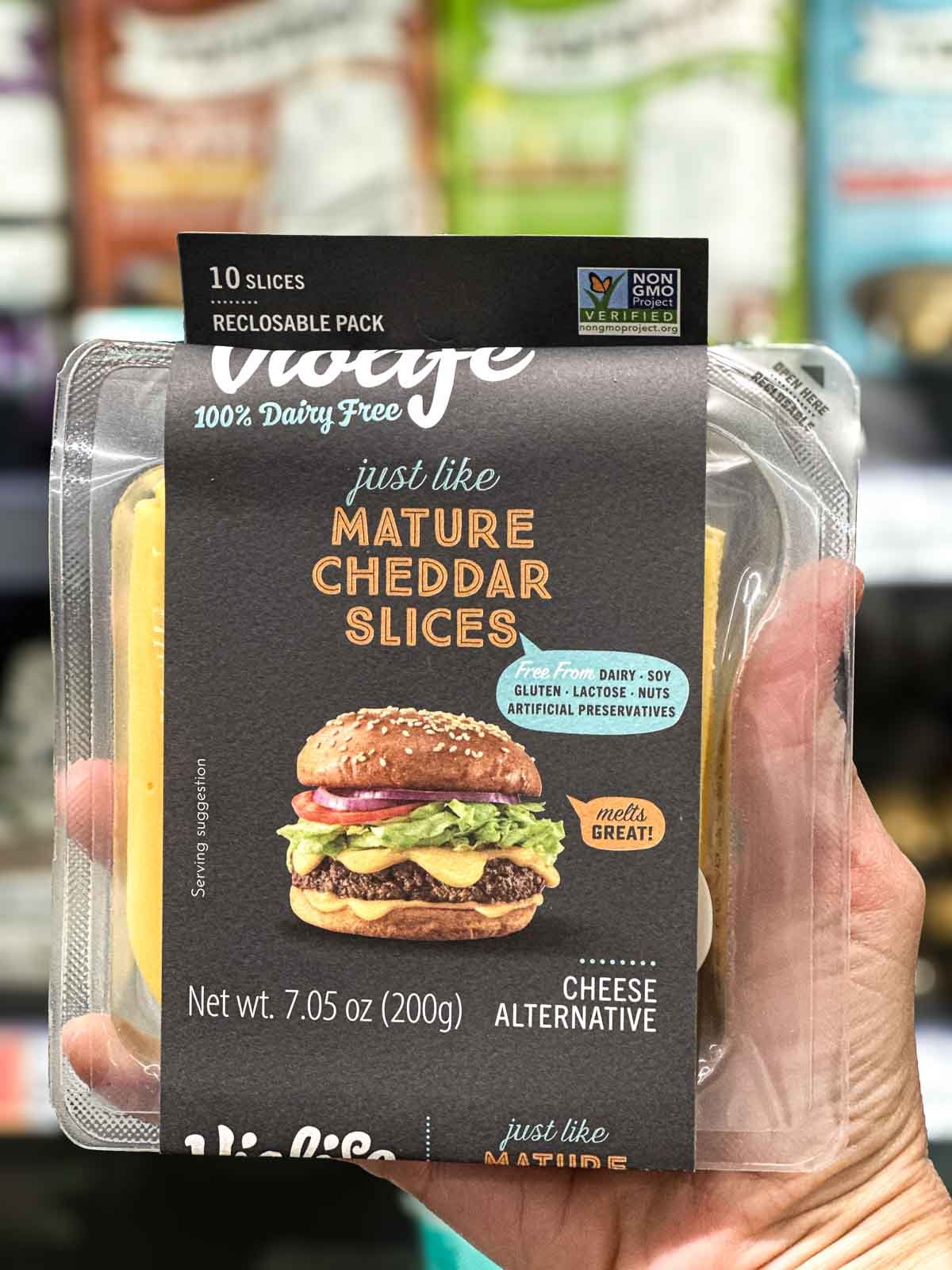 Violife vegan mature cheddar slices in reclosable pack held in hand, labeled 100% dairy free and melts great.