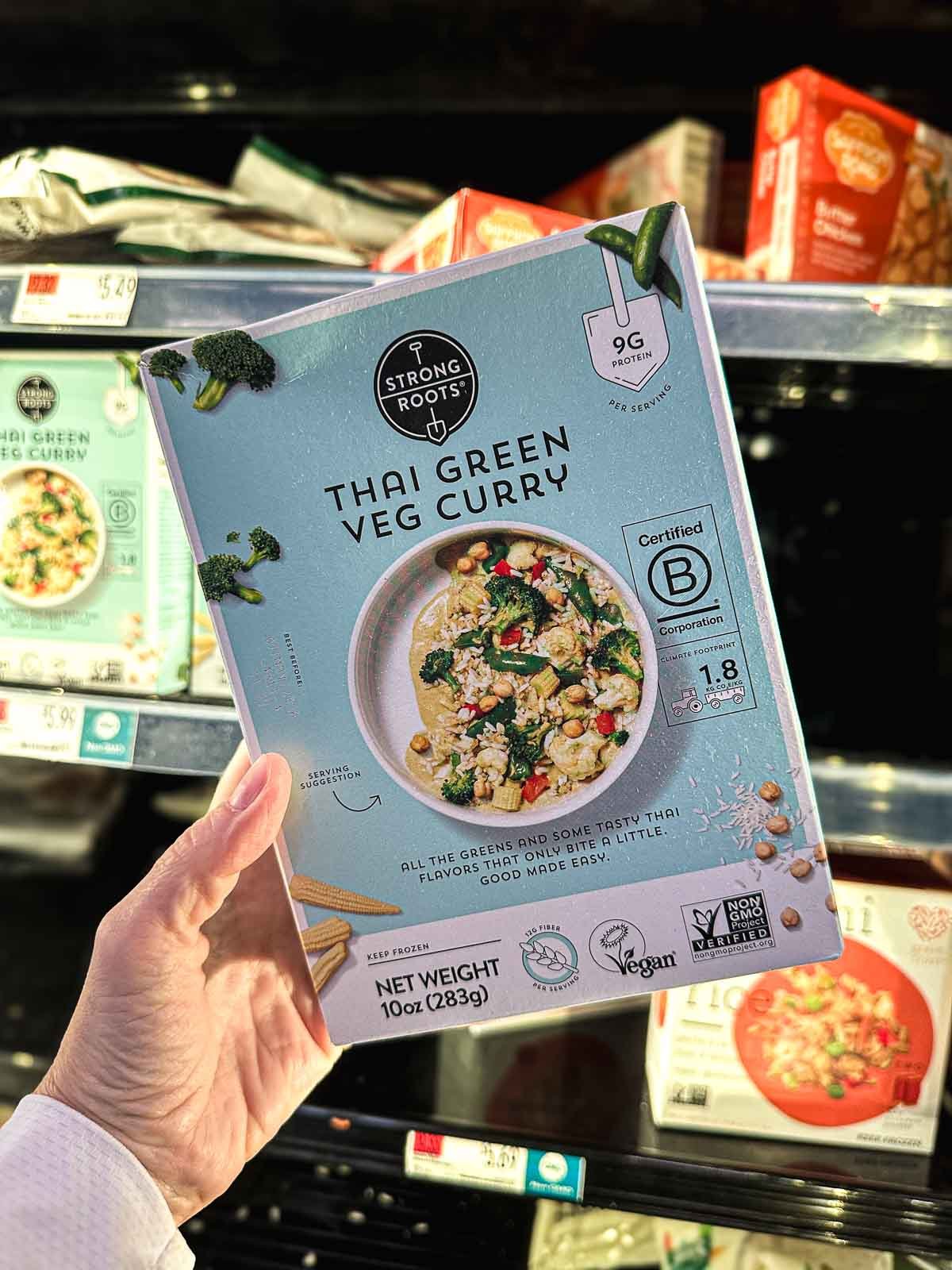 Box of Strong Roots Thai Green Veg Curry held in front of freezer case, labeled vegan and certified B Corp.