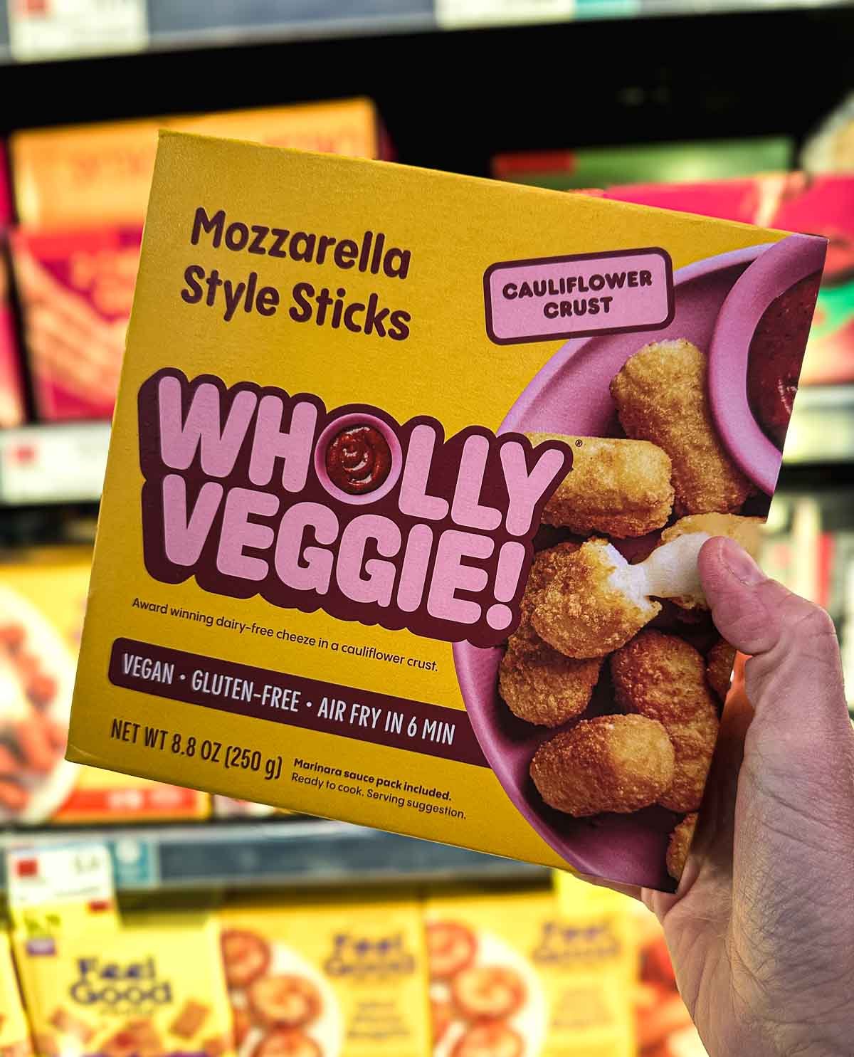 Wholly Veggie vegan mozzarella sticks in a yellow box, held up in the freezer section of a grocery store.