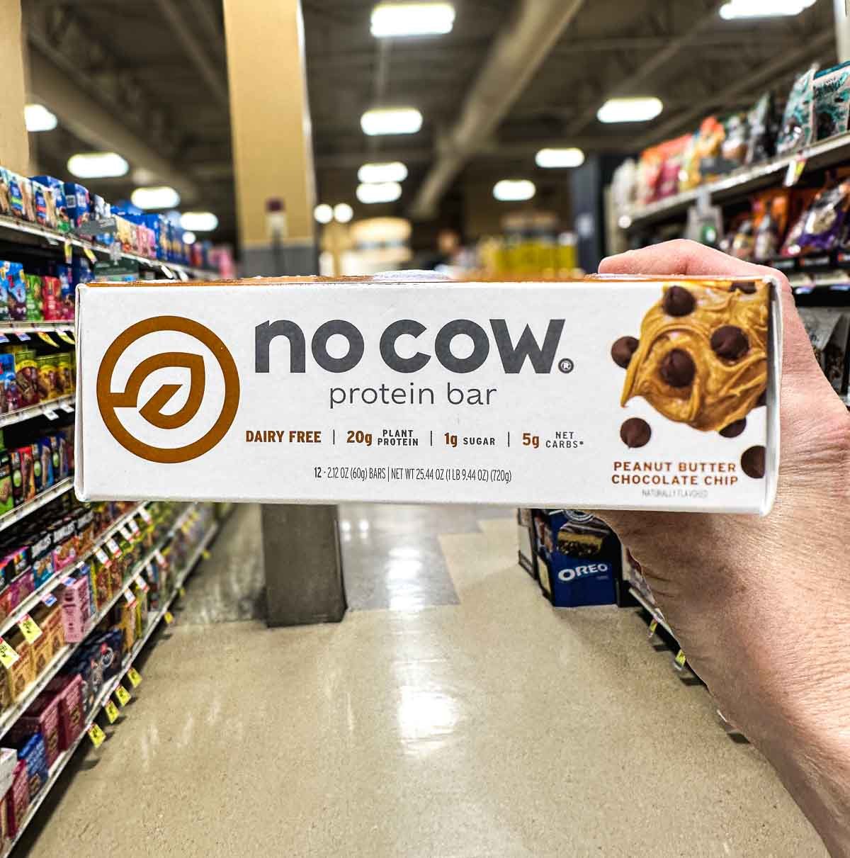 Hand holding No Cow vegan protein bar in peanut butter chocolate chip flavor, labeled 20g plant protein and dairy-free, shown in grocery store aisle.