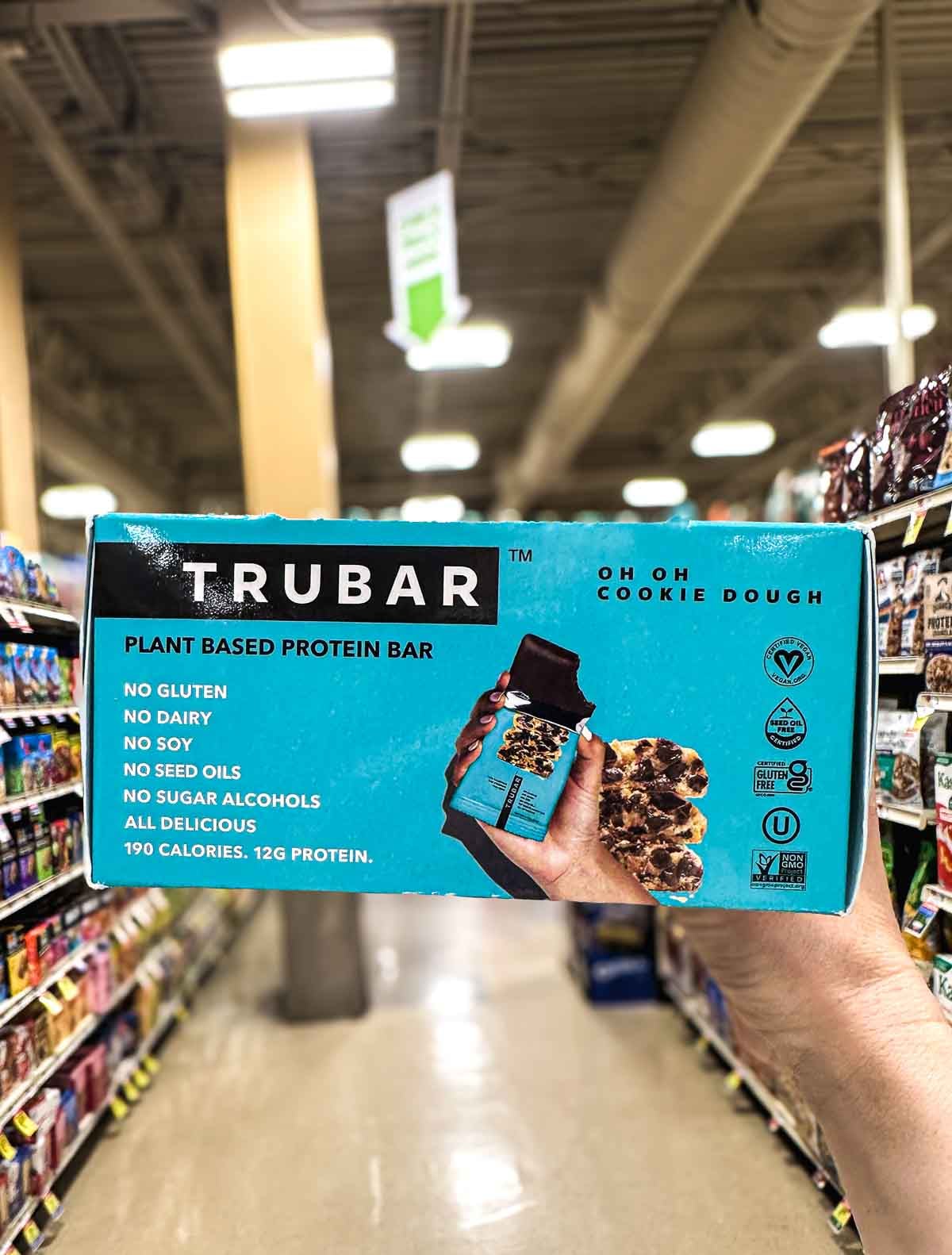 Hand holding TRUBAR Oh Oh Cookie Dough protein bars, plant-based and photographed in a grocery store aisle.