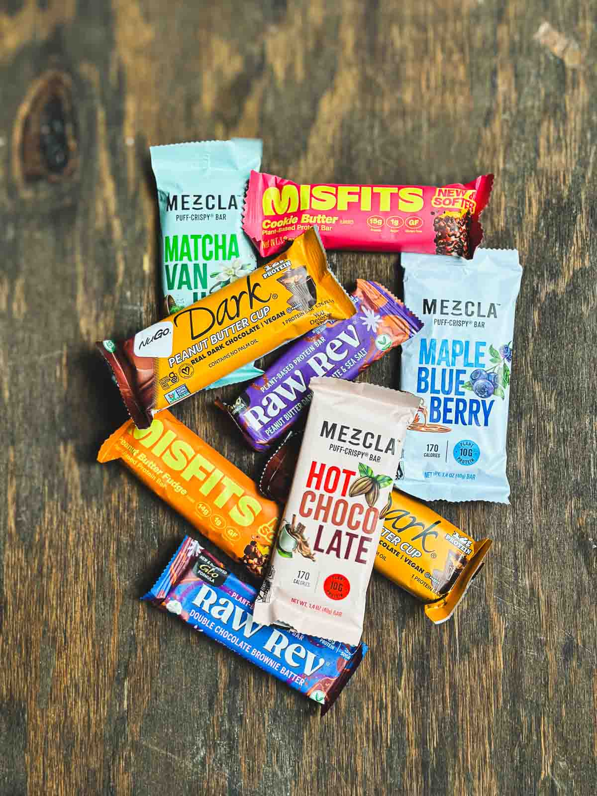 Assorted vegan protein bars including Raw Rev, Misfits, Mezcla, and NuGo stacked on a wood background.