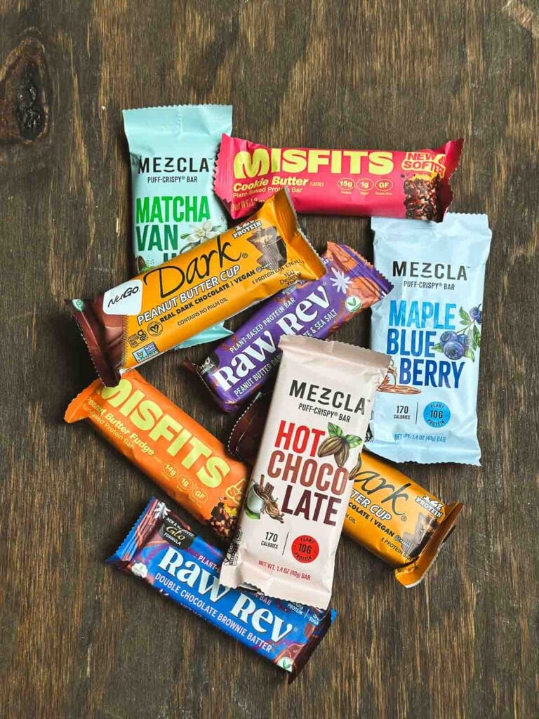 Top vegan protein bars like Mezcla, Misfits, Raw Rev, and NuGo displayed flat on a wooden surface.
