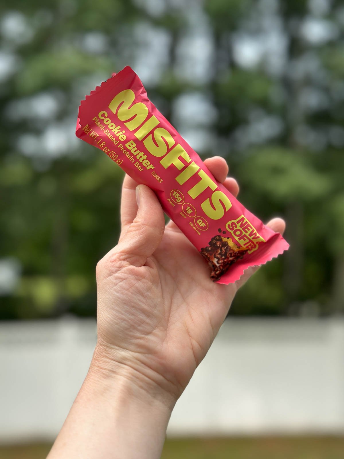 Close-up of a hand holding a pink Misfits Cookie Butter vegan protein bar with trees in the background.