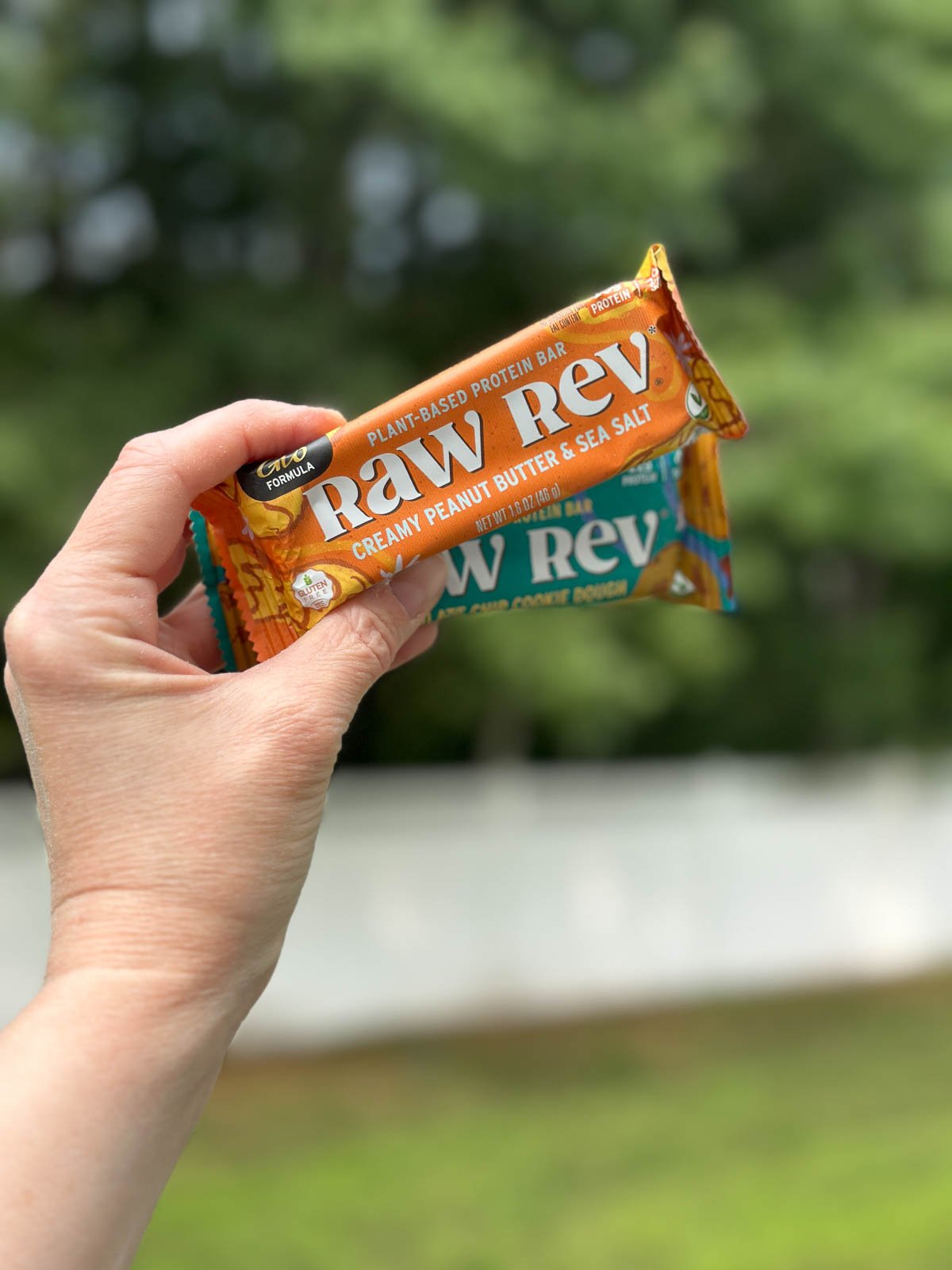 Hand holding two Raw Rev vegan protein bars, including Creamy Peanut Butter & Sea Salt, outdoors.