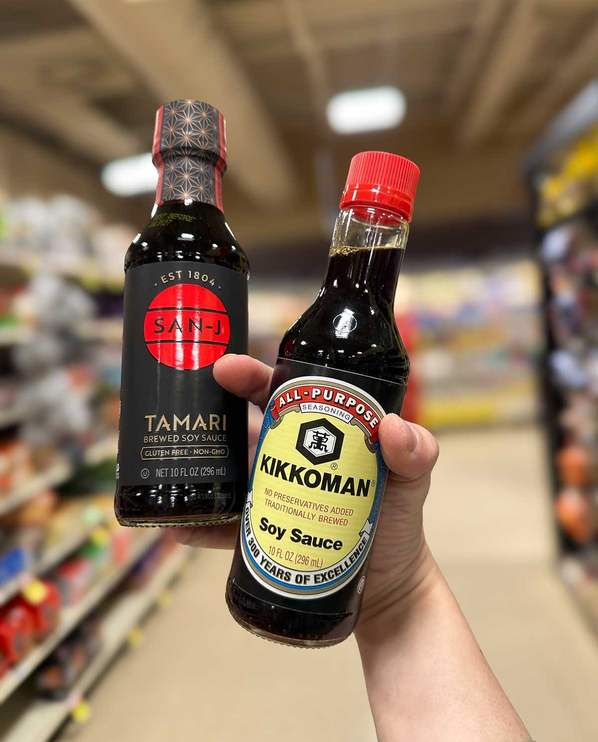 Hand holding bottles of San-J Tamari and Kikkoman Soy Sauce side by side on a grocery store aisle.