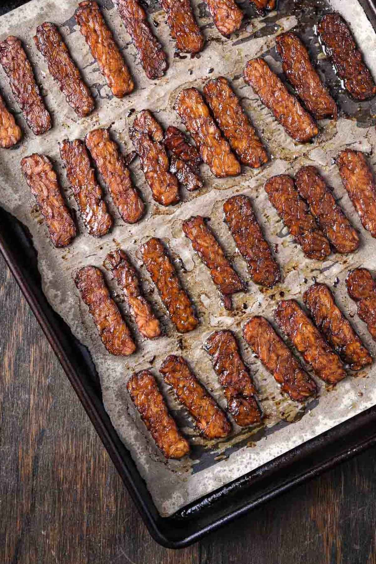 Crispy baked tempeh bacon on a parchment-lined baking sheet, caramelized and smoky from a maple-tamari marinade.