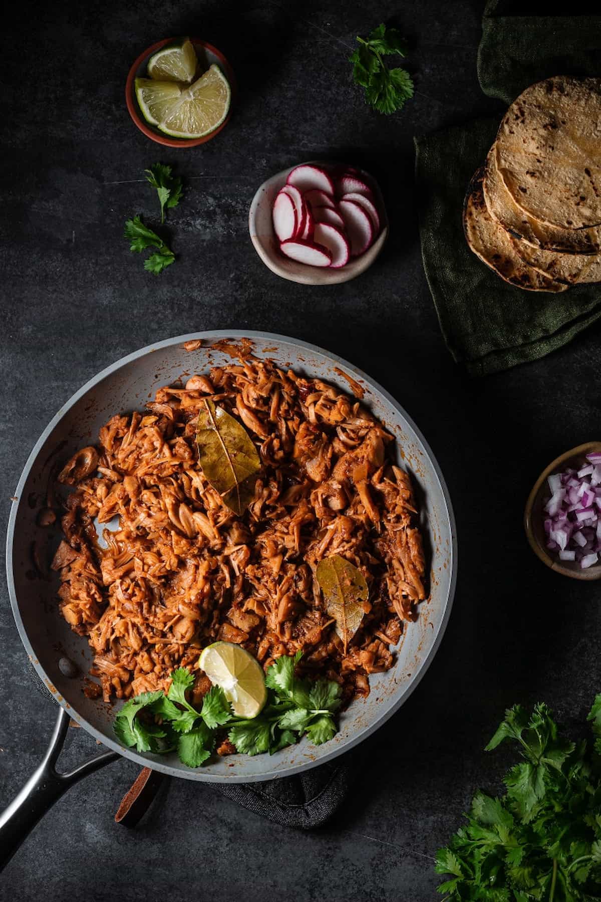 Jackfruit barbacoa simmered in chipotle sauce with bay leaves, lime, and cilantro in a skillet.