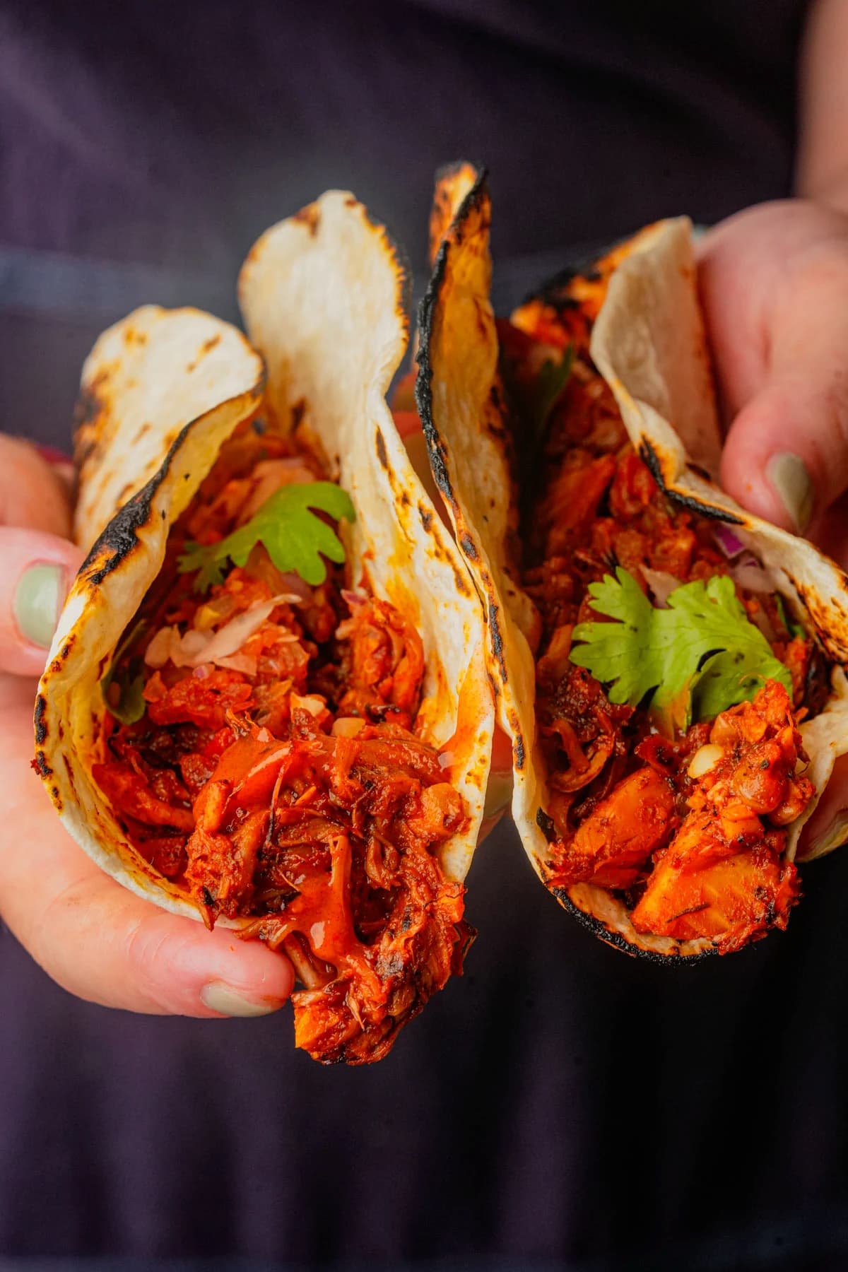 Close-up of jackfruit carnitas tacos with smoky red sauce and cilantro in charred tortillas.