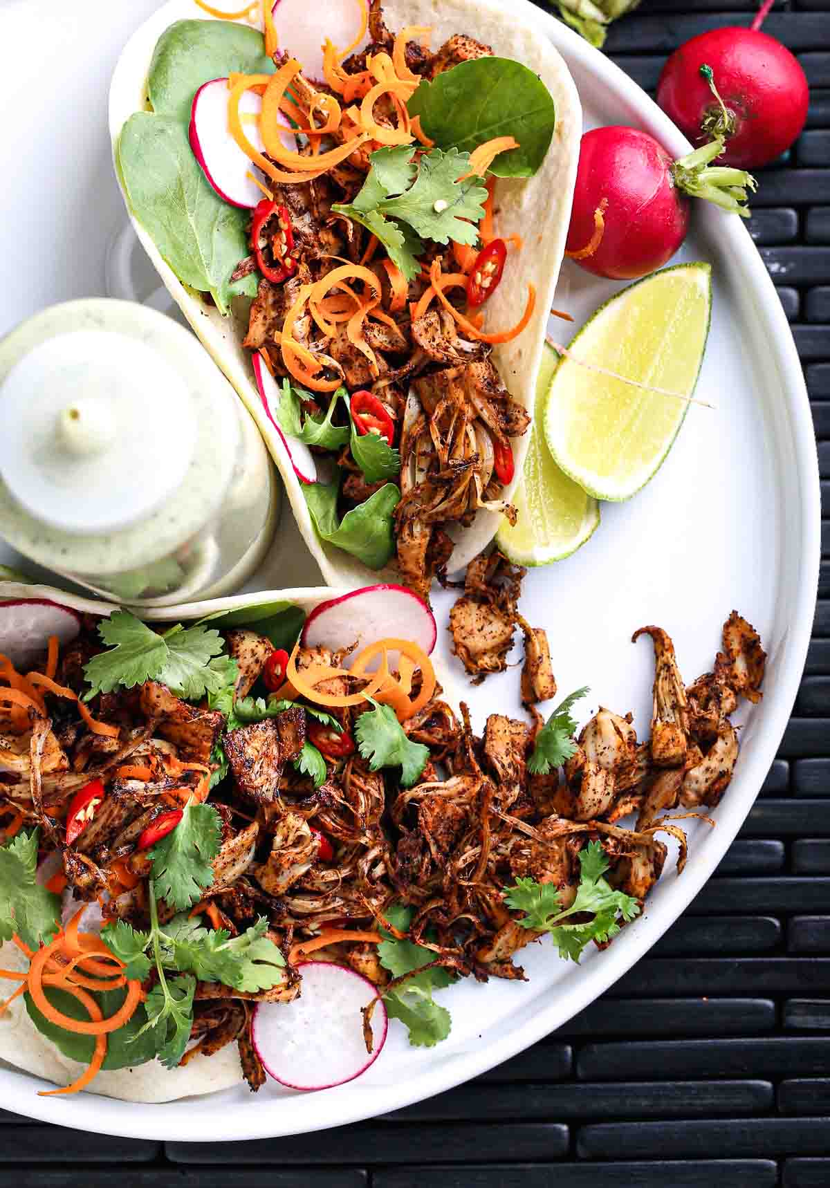 Spicy jackfruit tacos with shredded jackfruit, cilantro, radish, and lime on a white plate.