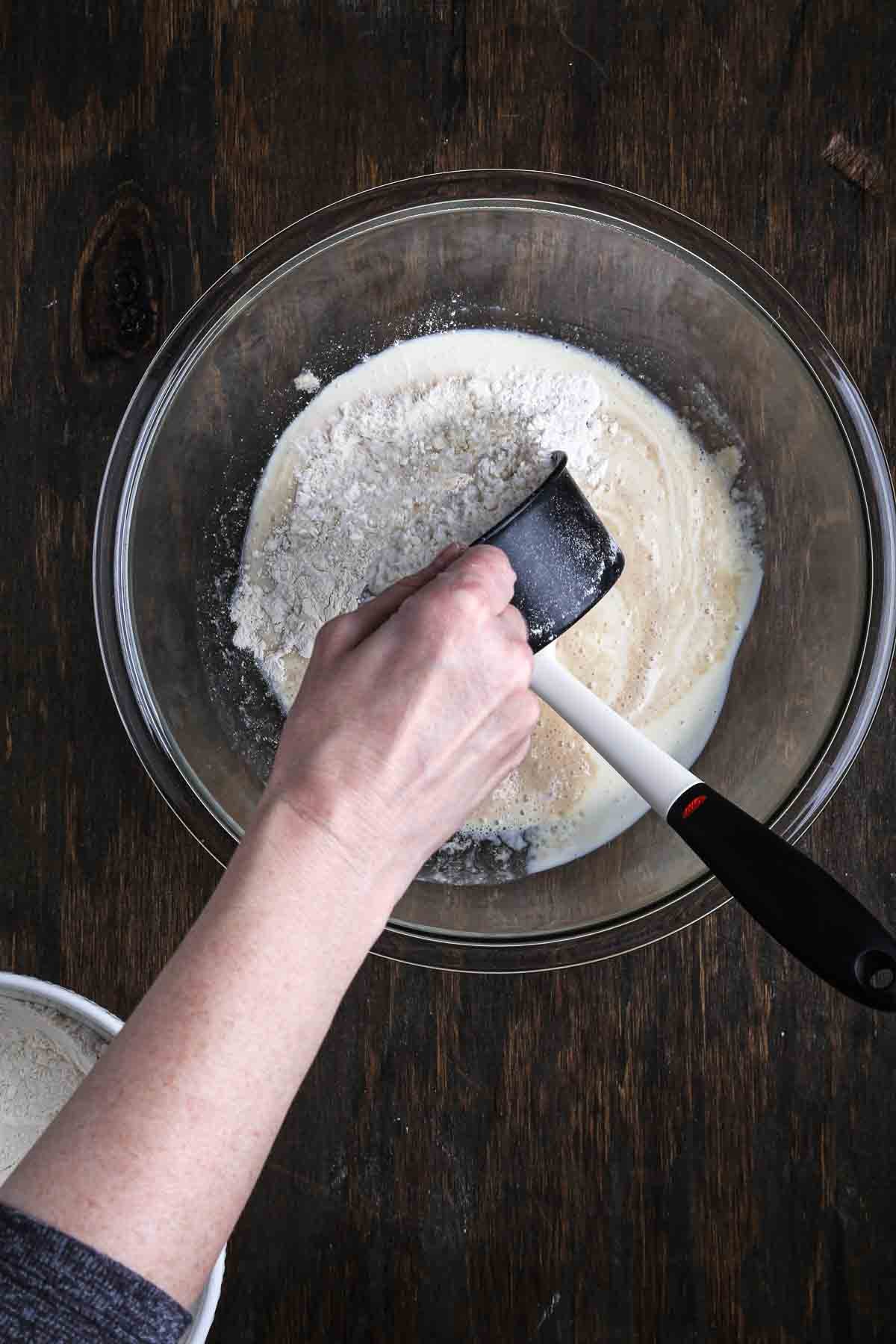 Hand adding a scoop of bread flour into the bowl of wet ingredients.