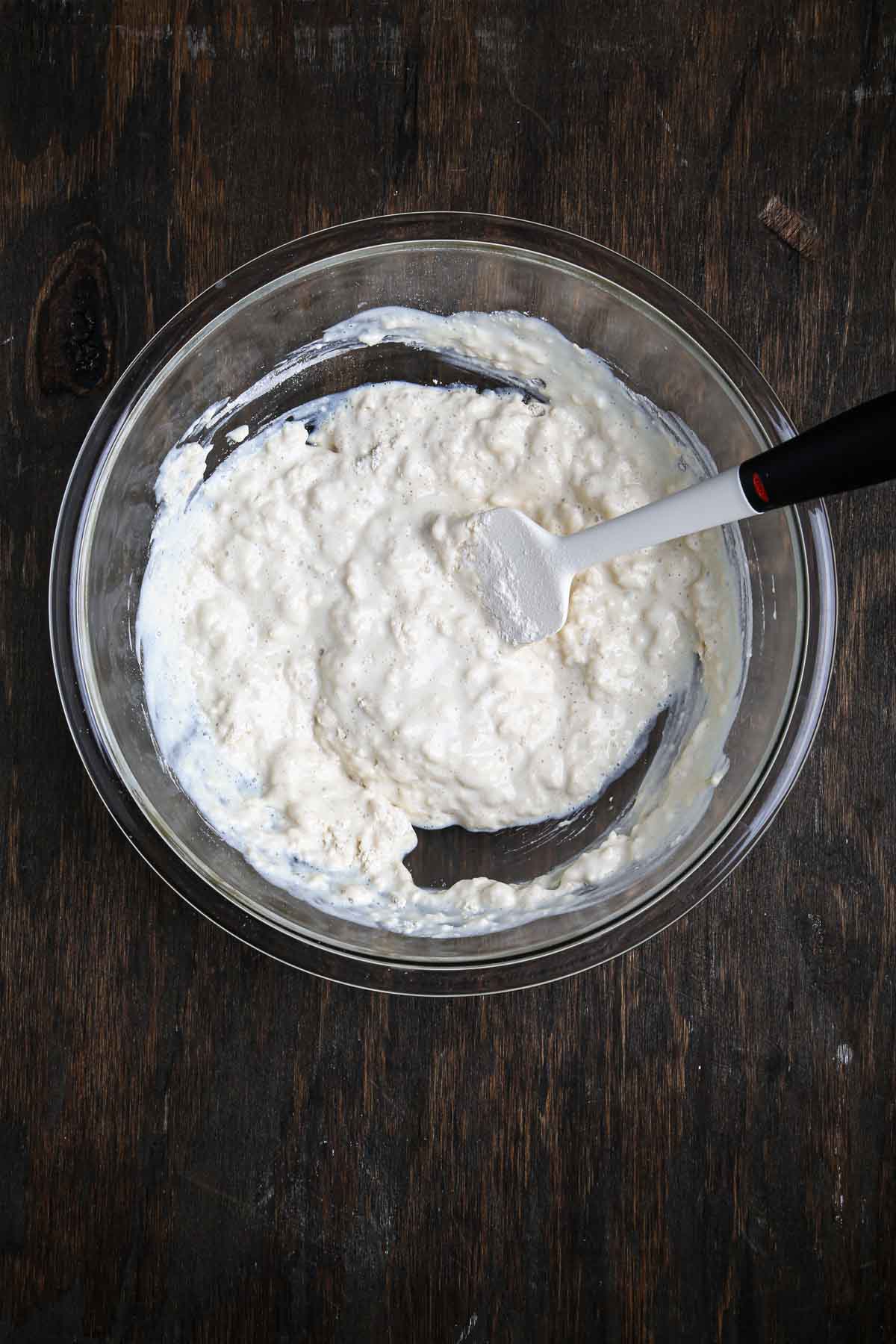 Partially mixed bread dough showing a loose, lumpy texture in a glass bowl.
