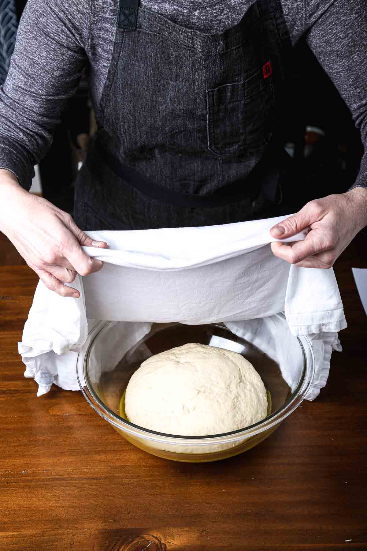 Risen bread dough in a glass bowl as a baker lifts the towel to check the first rise.