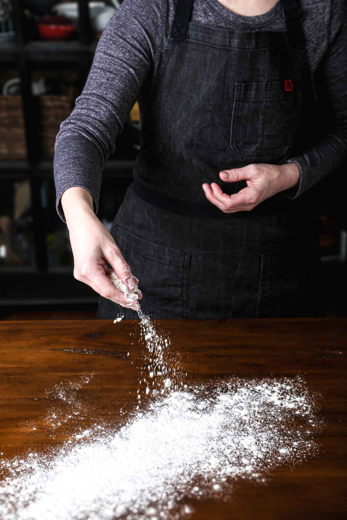 Hand sprinkling flour over a wooden surface to prepare for kneading.