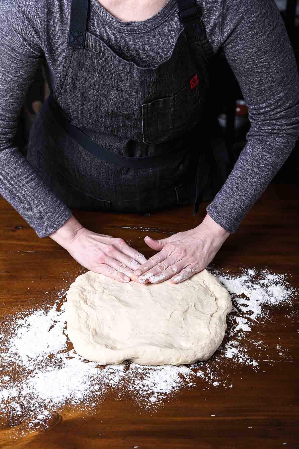 Hands pressing bread dough into a flat rectangle on a floured wooden surface.